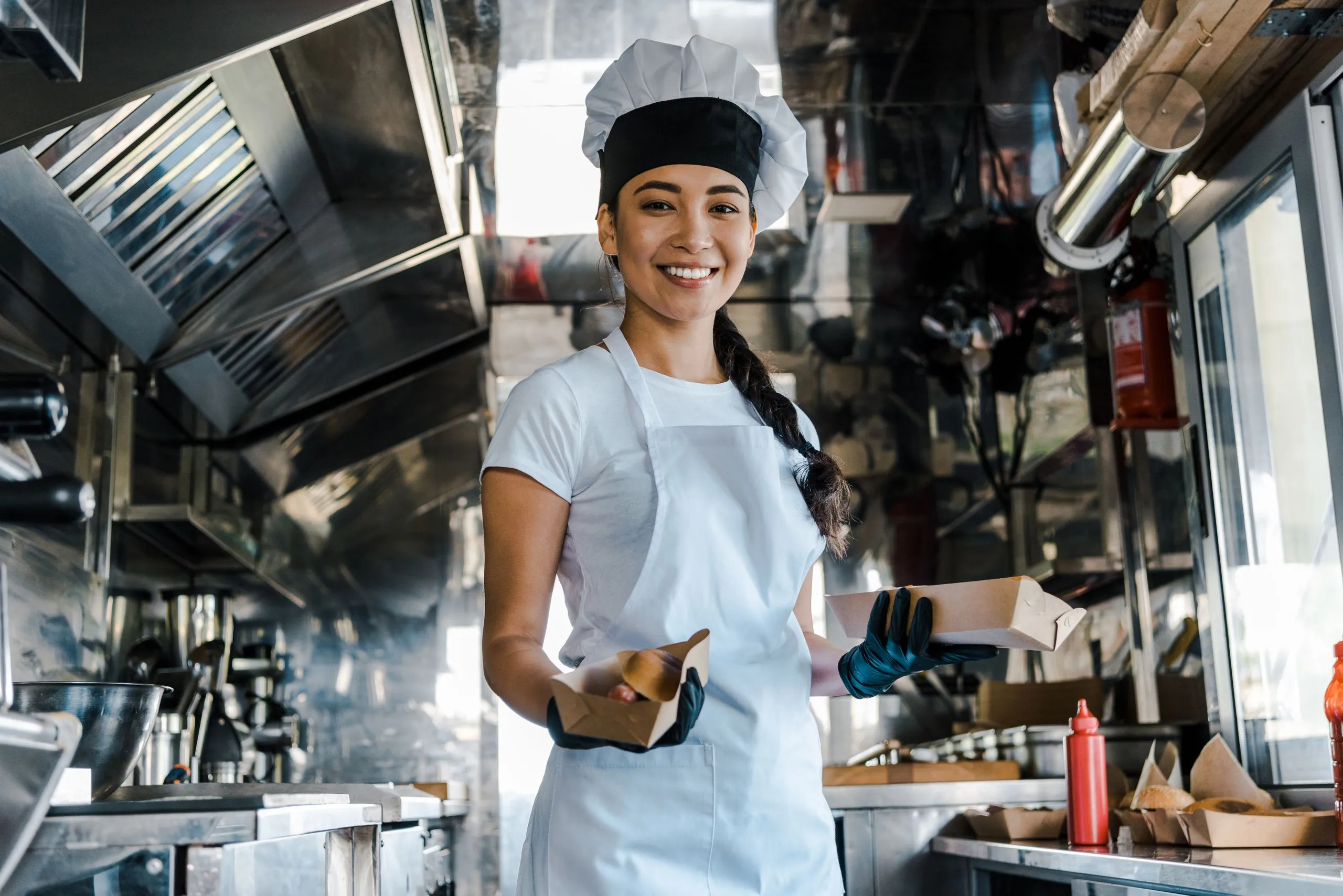 Cozinheira sorridente, de dólmã e touca, segura embalagens de comida em uma cozinha industrial